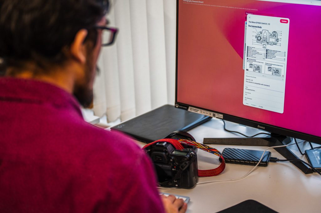 photograph of a man with a camera, sat in front of a computer looking at a diagram of. a camera
